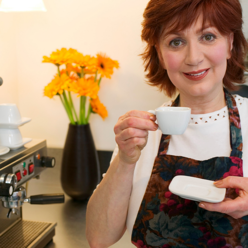 Woman holding a white cup in a kitchen setting with an espresso machine and flowers.