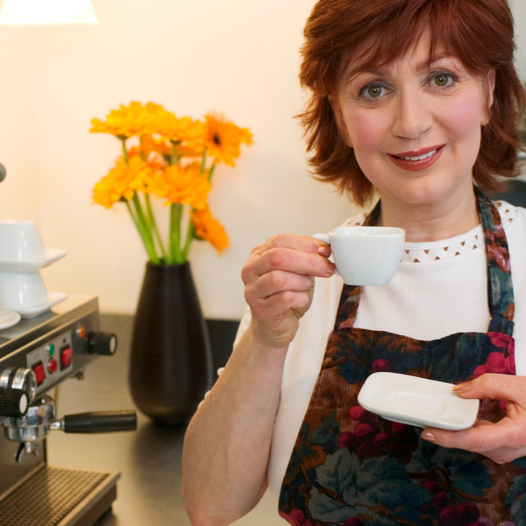 Woman holding a white cup in a kitchen setting with an espresso machine and flowers.