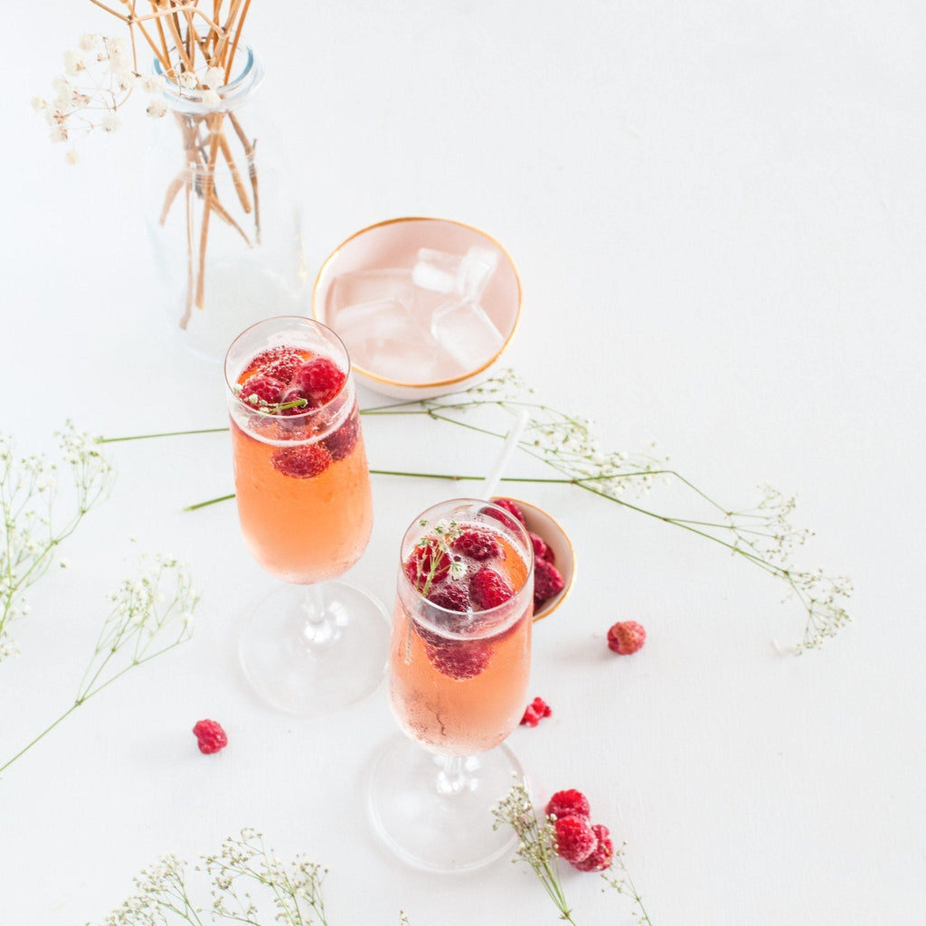 Two glasses of pink champagne with raspberries on a white surface with flowers.