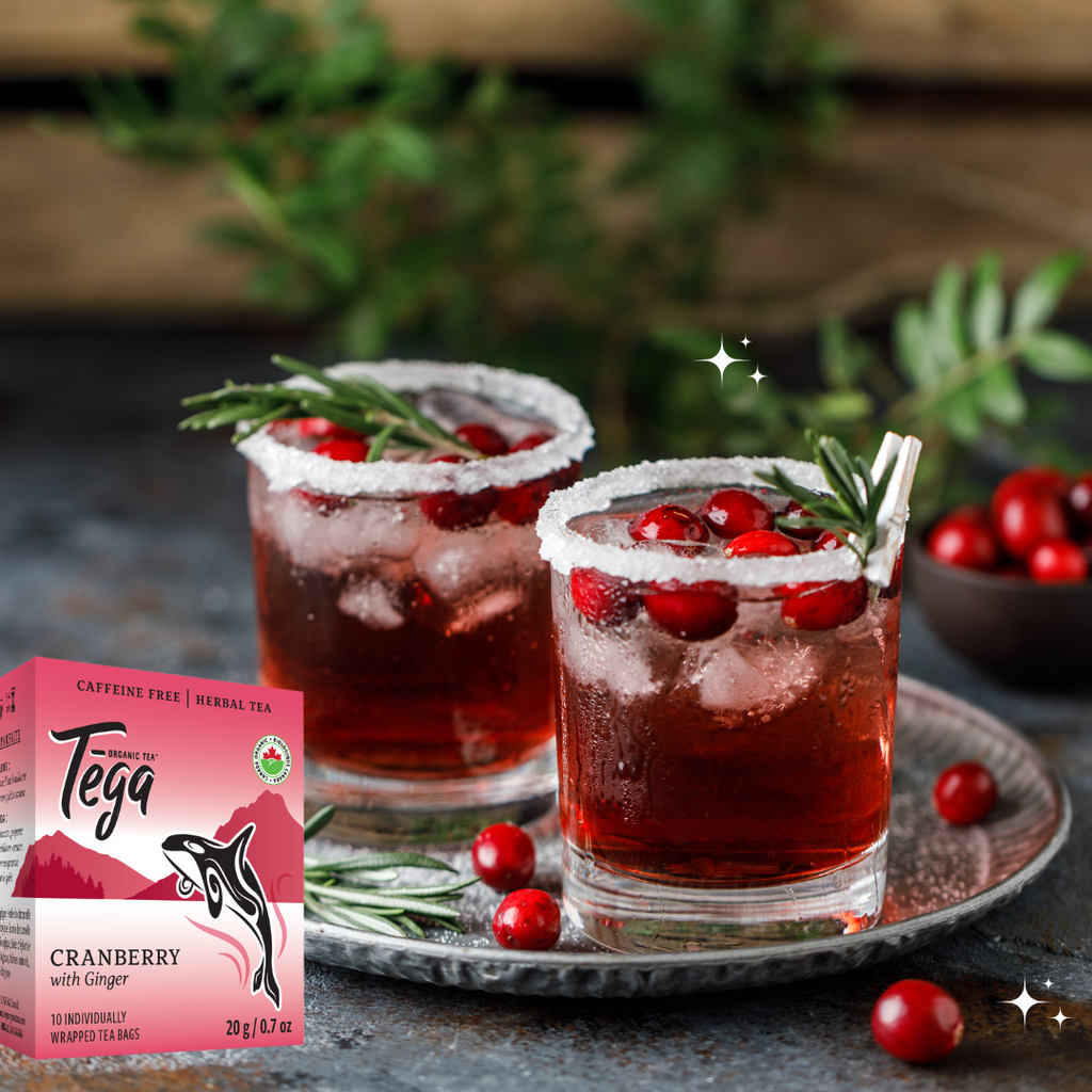 Two glasses of cranberry juice with ice and garnishes, next to a box of Tega Cranberry with Ginger tea.