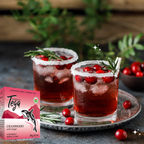 Two glasses of cranberry juice with ice and garnishes, next to a box of Tega Cranberry with Ginger tea.