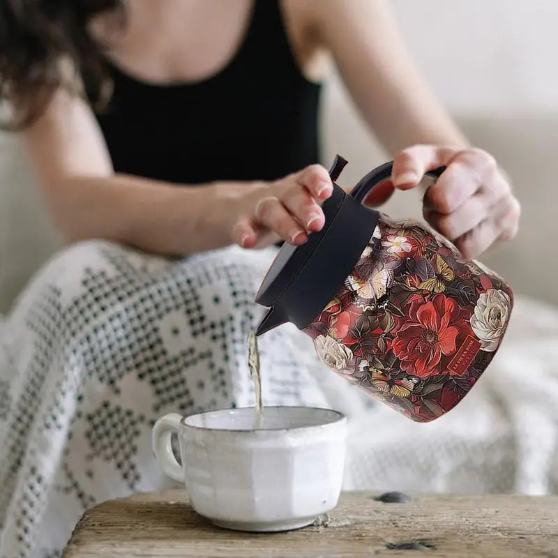 Person pouring tea from a floral-patterned teapot into a white mug.