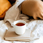 White mug with tea on a stack of books under a beige blanket and pillows.