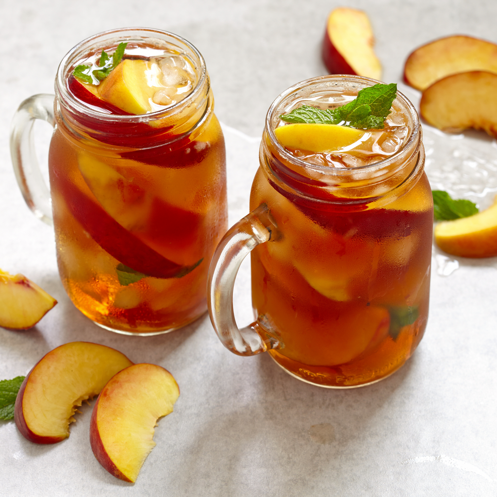 Two mason jars filled with peach iced tea on a light surface with sliced peaches around.